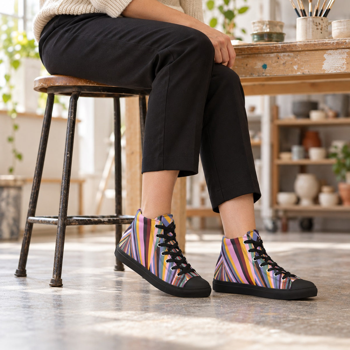 Woman seated in an art studio wearing Dynamic Radiating Stripe high-top sneakers with black soles and cropped black trousers.