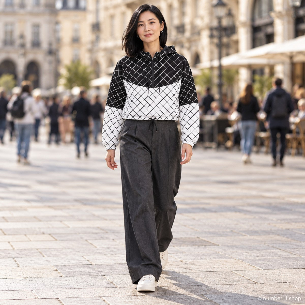 Woman wearing a black and white grid cropped windbreaker with charcoal wide-leg trousers and white sneakers in a busy city square.