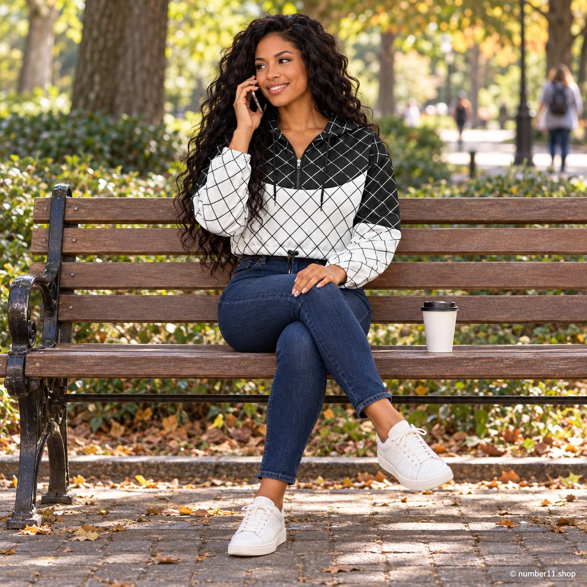 Woman wearing a black and white grid cropped windbreaker and jeans, sitting cross-legged on a park bench while talking on her phone, with coffee beside her.