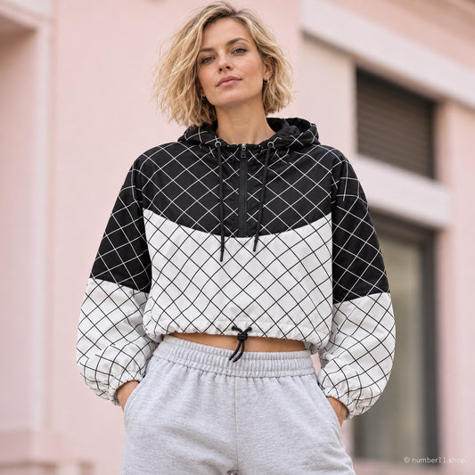 Woman with a blonde bob wearing a graphic black and white grid cropped windbreaker with grey joggers against a pale pink urban wall.